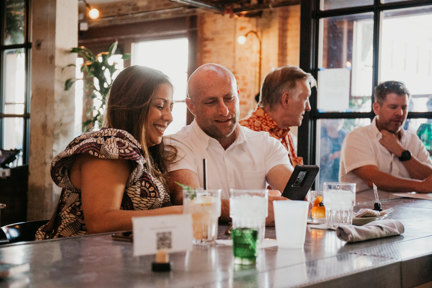 Image of a woman and man sitting at a bar, they're smiling and looking at a phone with drinks in front of them